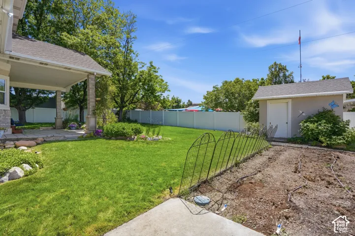 Expansive backyard featuring a stone patio, manicured lawn, dedicated garden area, white privacy fencing, and a detached storage shed