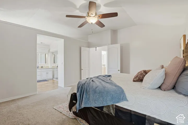 Vaulted ceiling with a ceiling fan light fixture, carpeted flooring, and an open doorway leading to a bathroom featuring a double vanity with white cabinetry and framed mirrors
