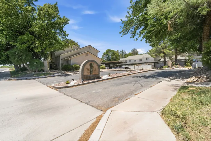 Community entrance featuring a stone monument sign, mature trees, paved roadways, and residential structures