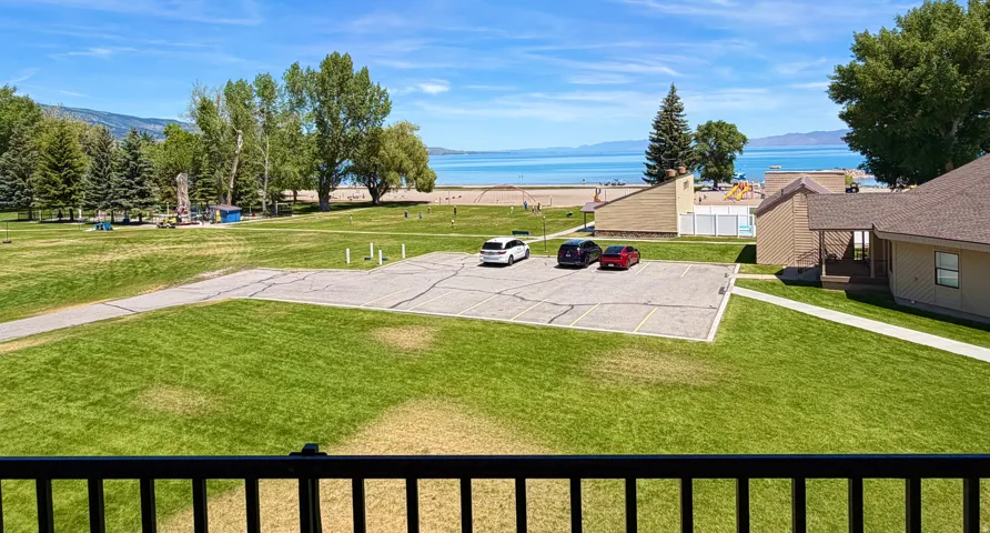 Expansive green space with mature trees, paved pathways, and a parking area, all backing directly onto a sandy beach and a large body of water