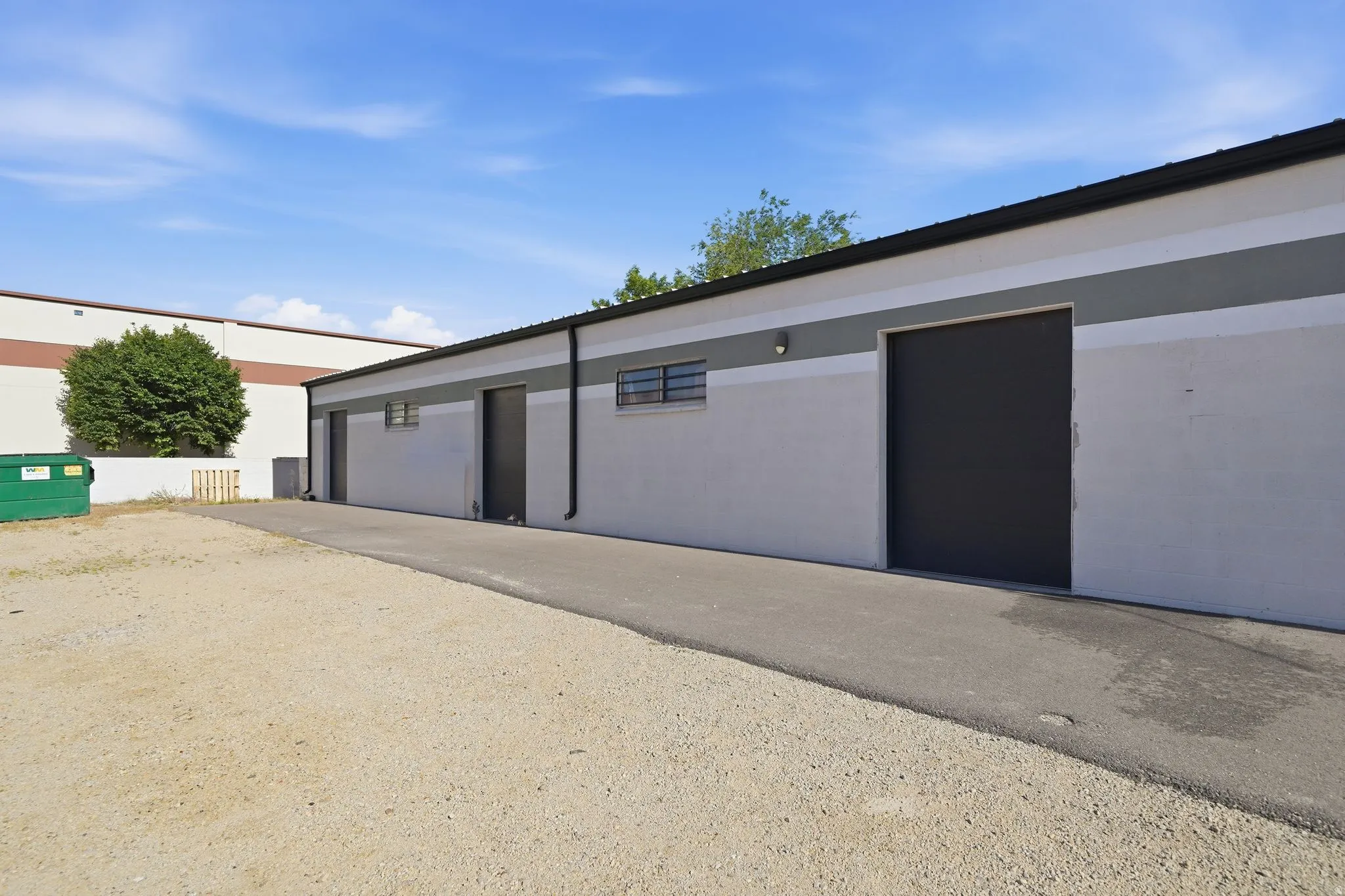Industrial building featuring a light gray exterior with dark gray and white horizontal banding, multiple roll-up doors, and wall-mounted exterior lighting