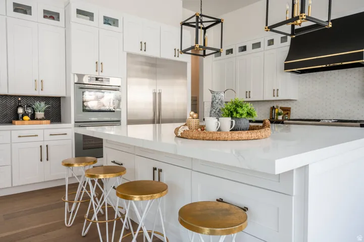 Bright kitchen with a large white island, white shaker cabinetry, stainless steel appliances including a built-in oven and refrigerator, a black and gold range hood, and wood-finish flooring