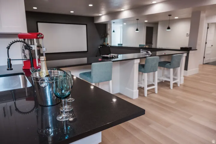 Spacious entertainment area featuring a projection screen, an expansive L-shaped bar with seating, black granite countertops, and light wood-finish flooring