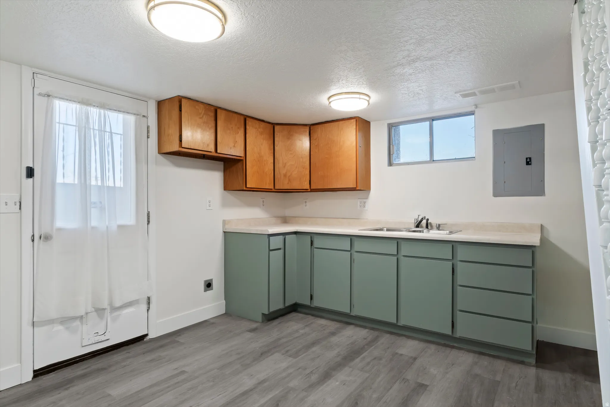 Utility kitchen area featuring grey wood-finish flooring, a stainless steel double basin sink, light green lower cabinetry, and natural wood upper cabinetry
