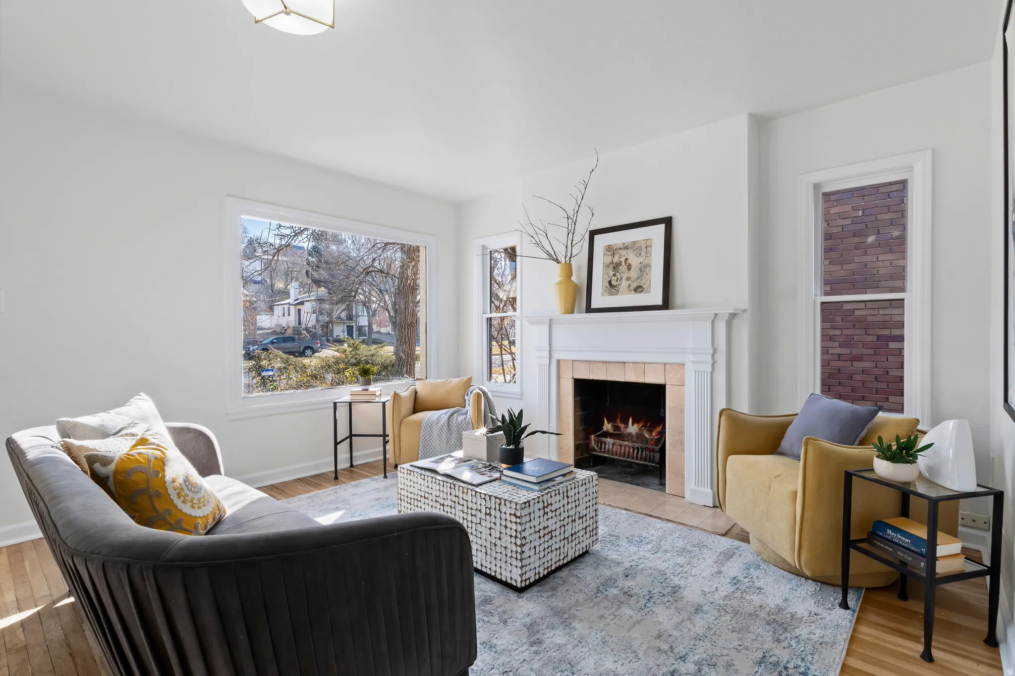 Bright living area featuring wood-finish flooring, a decorative fireplace with a white mantel, and large windows providing abundant natural light