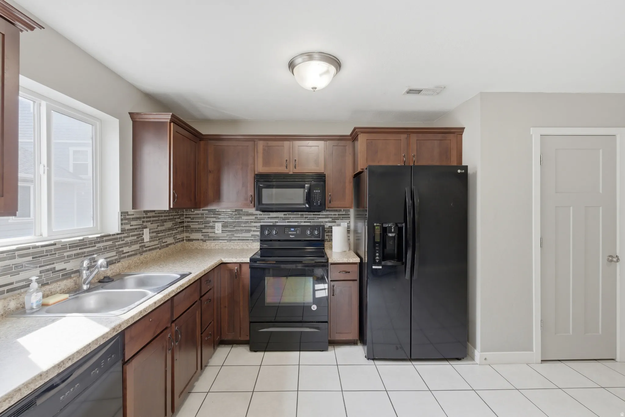 Kitchen featuring dark wood cabinetry, a mosaic tile backsplash, black appliances, a double basin stainless steel sink, and light-toned countertops