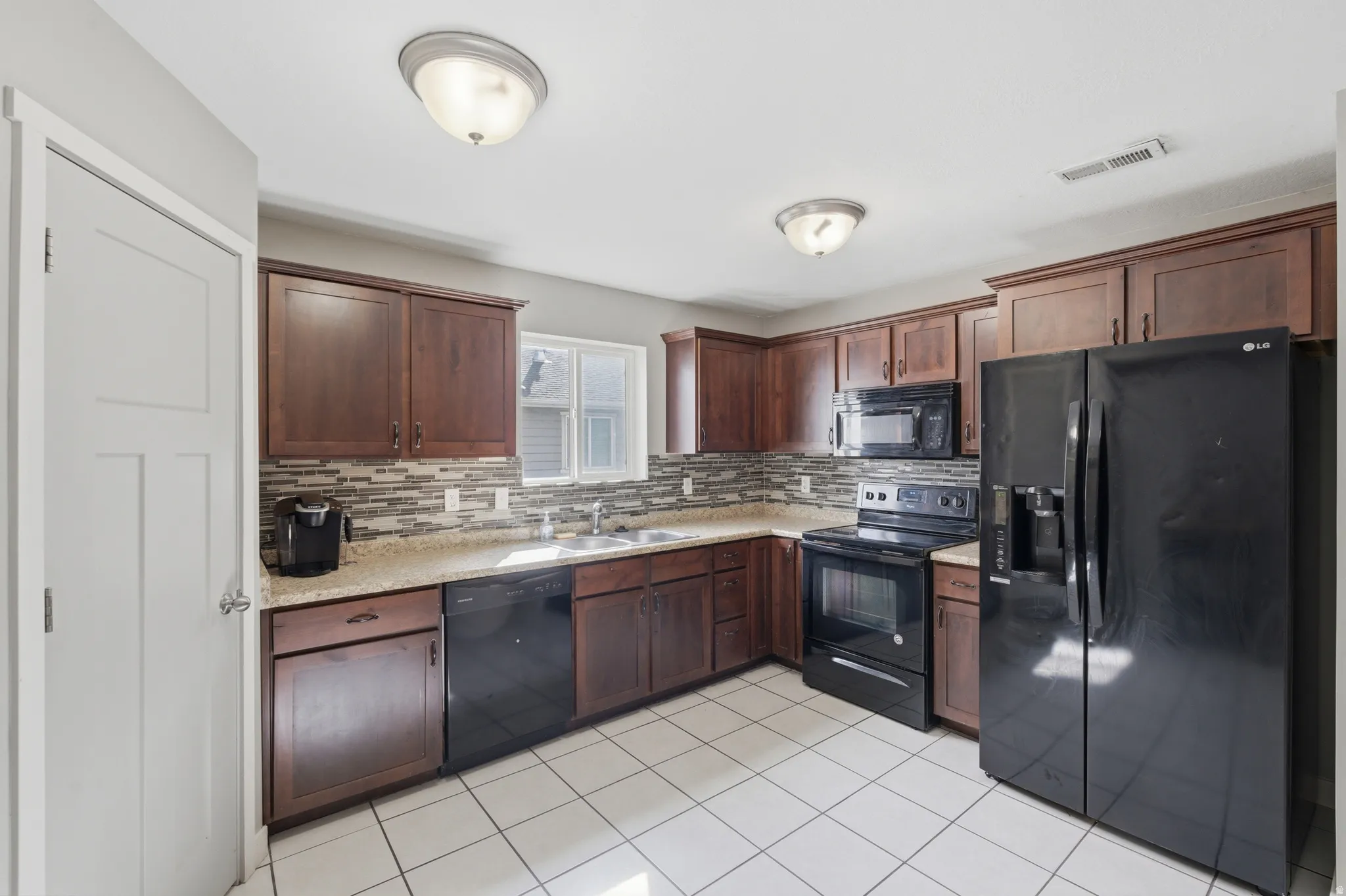 Kitchen featuring white tile flooring, dark wood cabinetry, light-toned countertops, a multi-tone tile backsplash, and black appliances