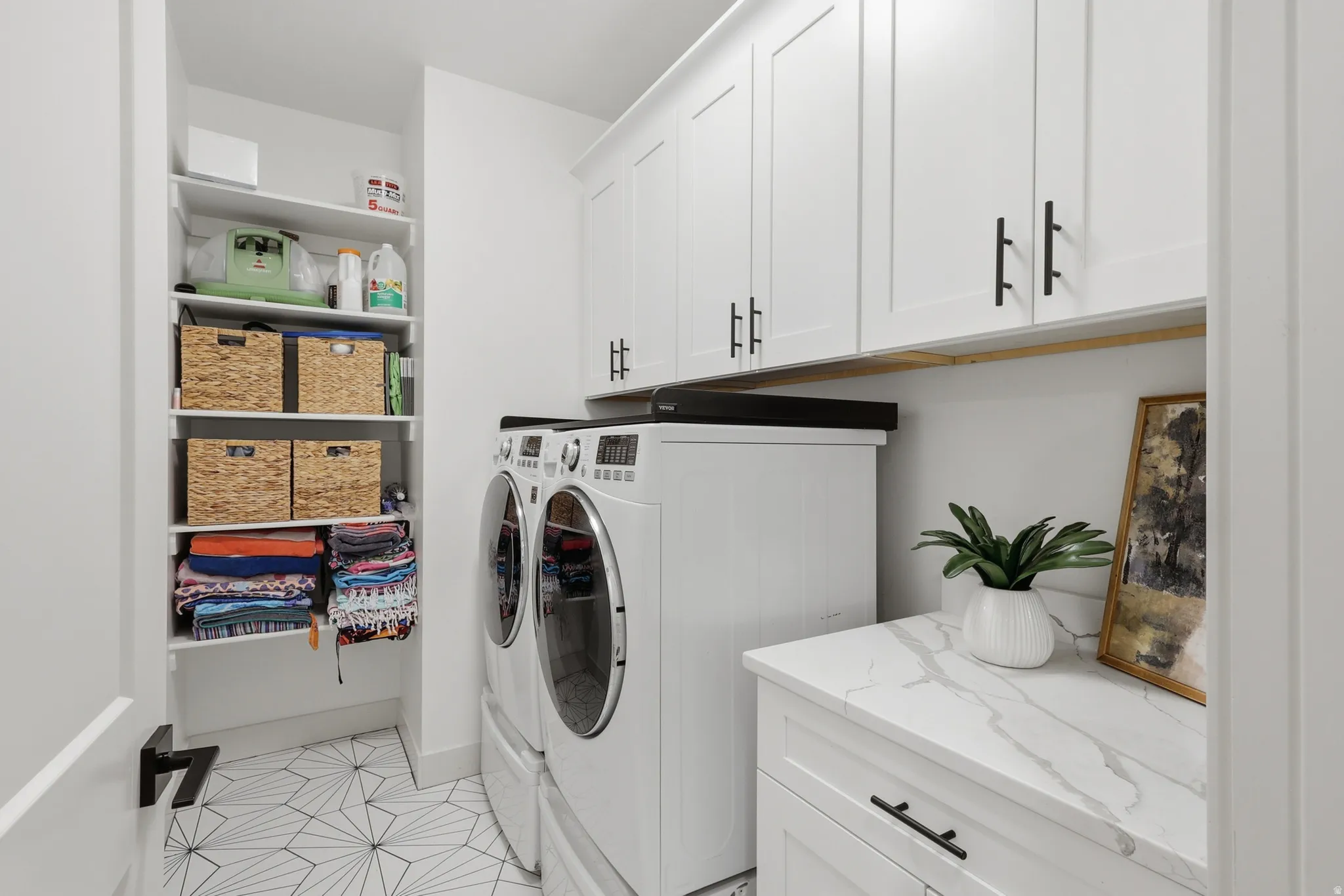 Dedicated laundry space featuring stacked appliances, overhead white shaker cabinetry with black hardware, an integrated counter with a marble-finish surface, and a built-in shelving unit