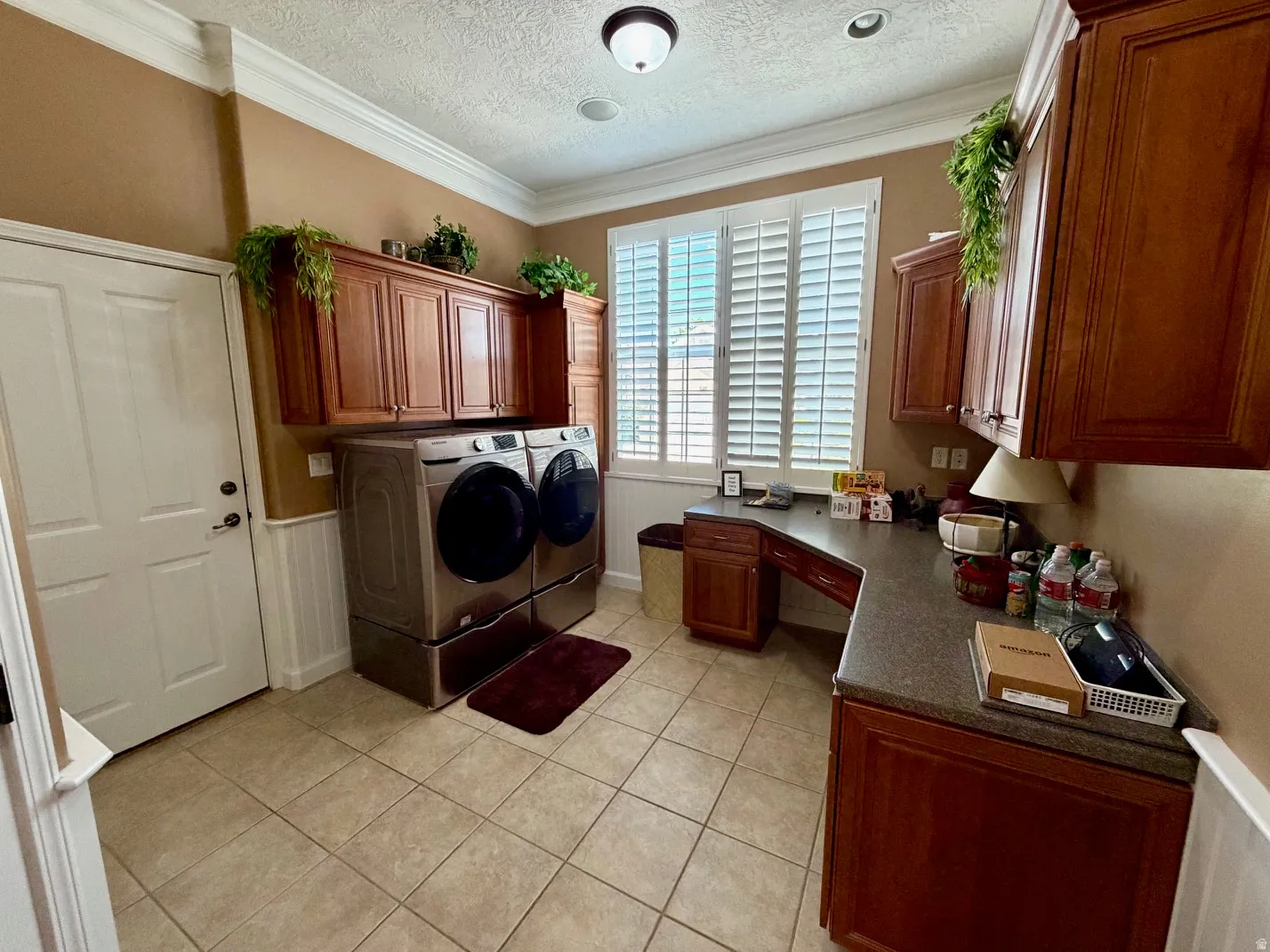 Spacious utility room featuring extensive built-in cabinetry, durable tile flooring, a custom countertop workspace, crown molding, and plantation shutters