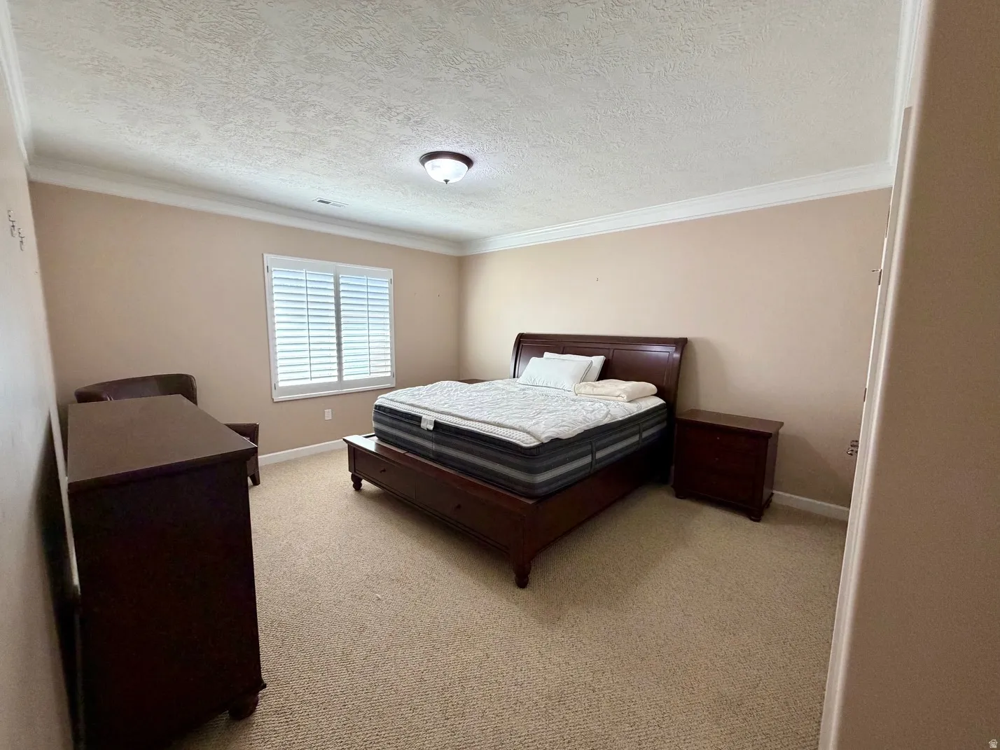 Spacious room with a neutral color palette, featuring textured ceilings, white crown molding, and a large window with white plantation shutters