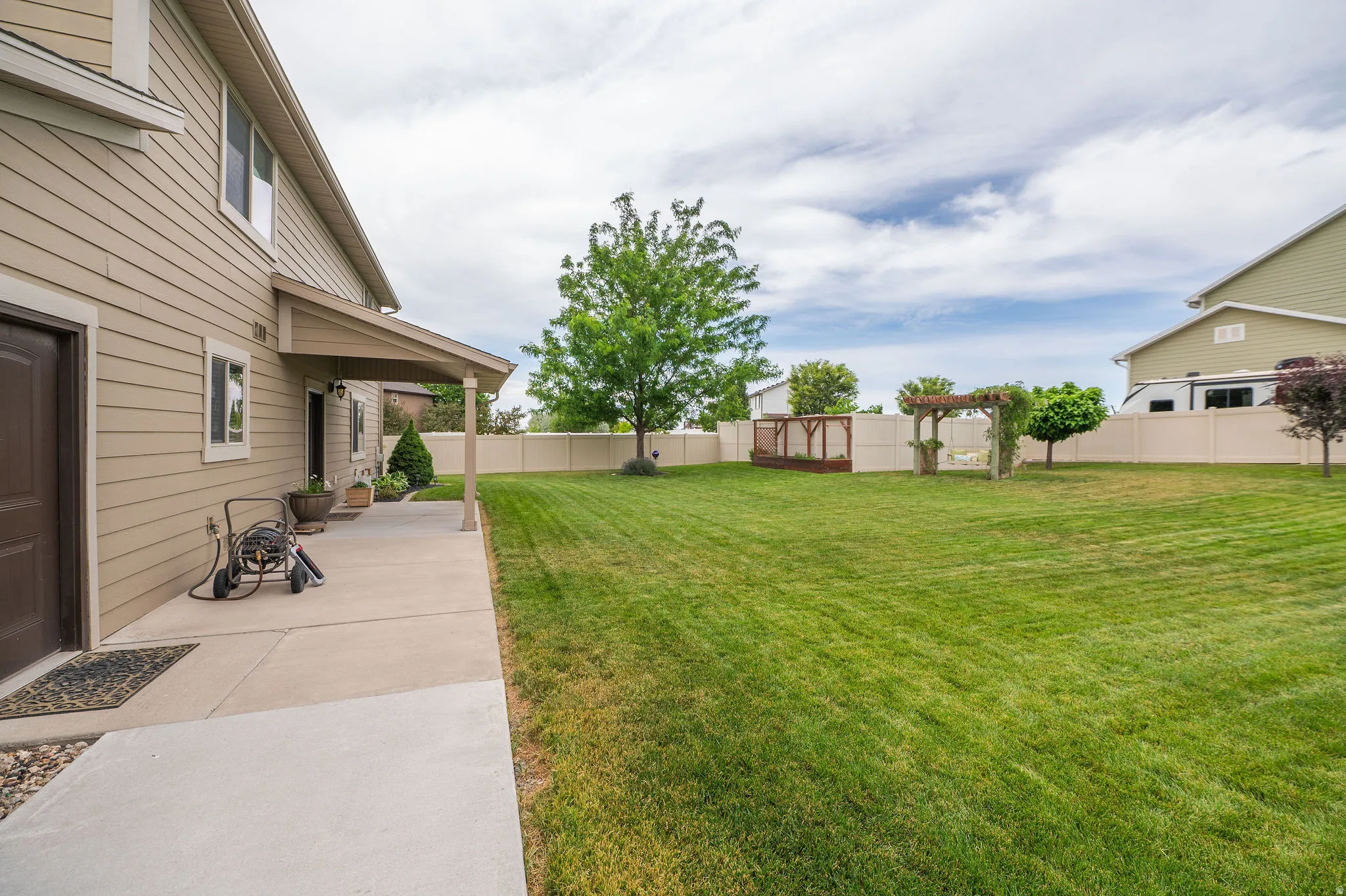 Expansive backyard featuring a manicured lawn, a covered concrete patio, a wooden pergola, a small raised deck structure, and full perimeter fencing