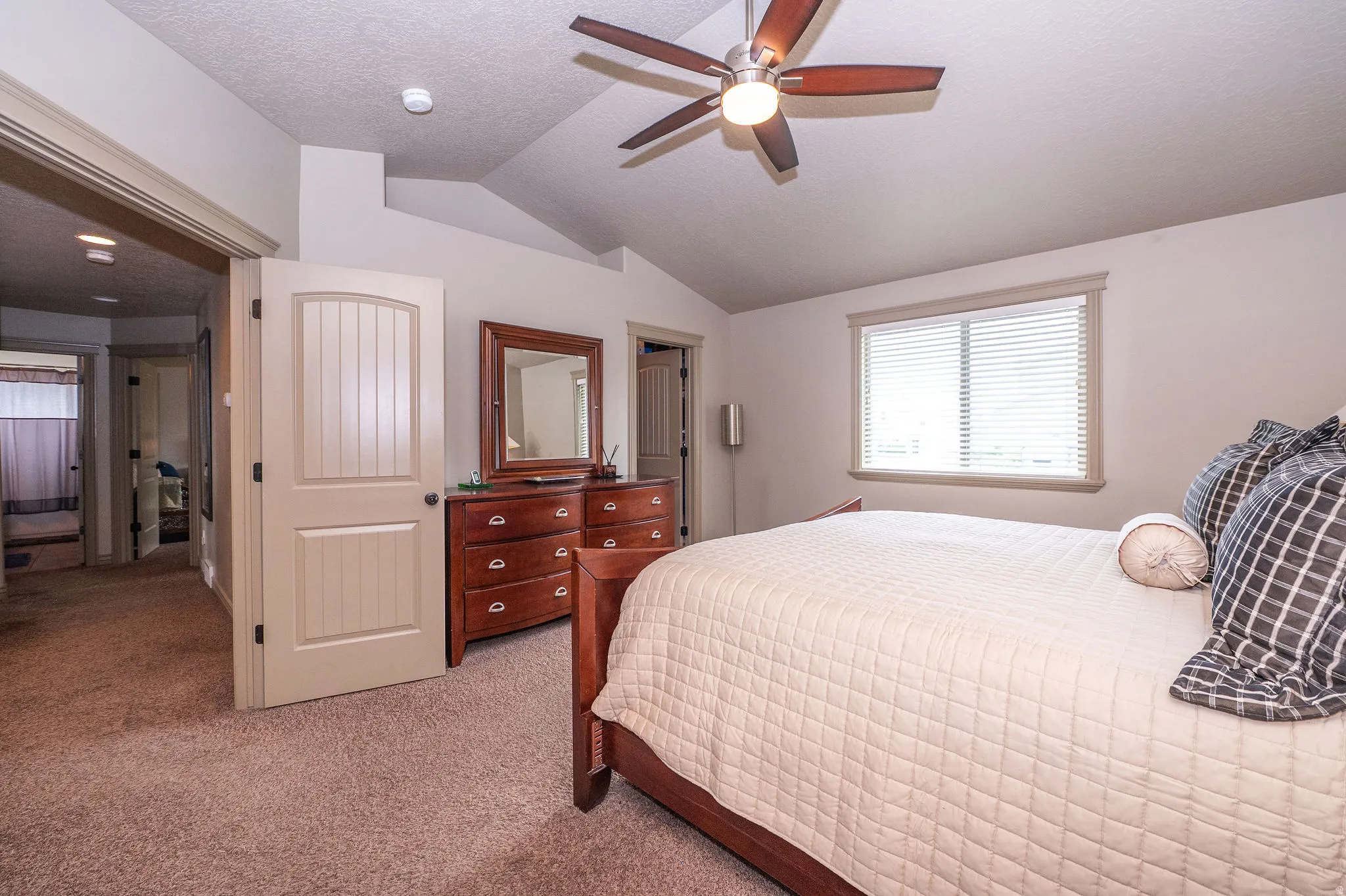 Bedroom featuring vaulted ceilings, a ceiling fan with light fixture, light-colored carpet, a window with horizontal blinds, and a paneled interior door