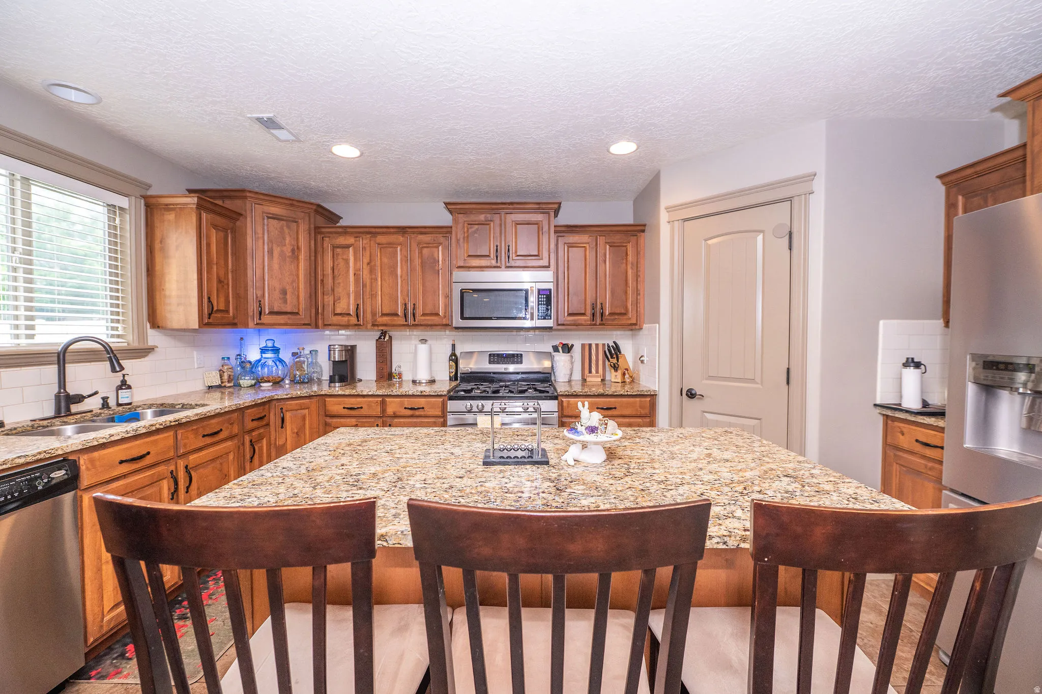 Kitchen featuring a spacious island with granite countertops, wood-finish cabinetry, stainless steel appliances, a tile backsplash, and recessed lighting