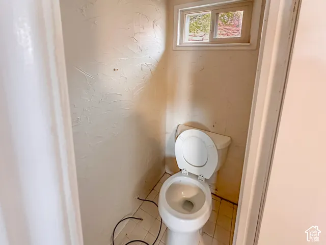 Compact water closet featuring a standard toilet, square tiled flooring, and a single-pane window