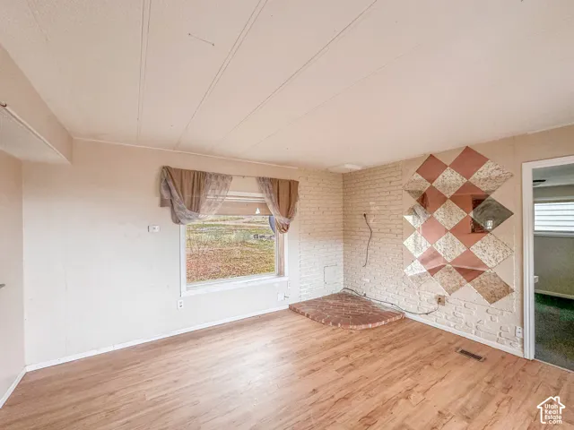 Spacious room featuring wood-finish flooring, a large window, painted white brick wall, and a raised brick hearth