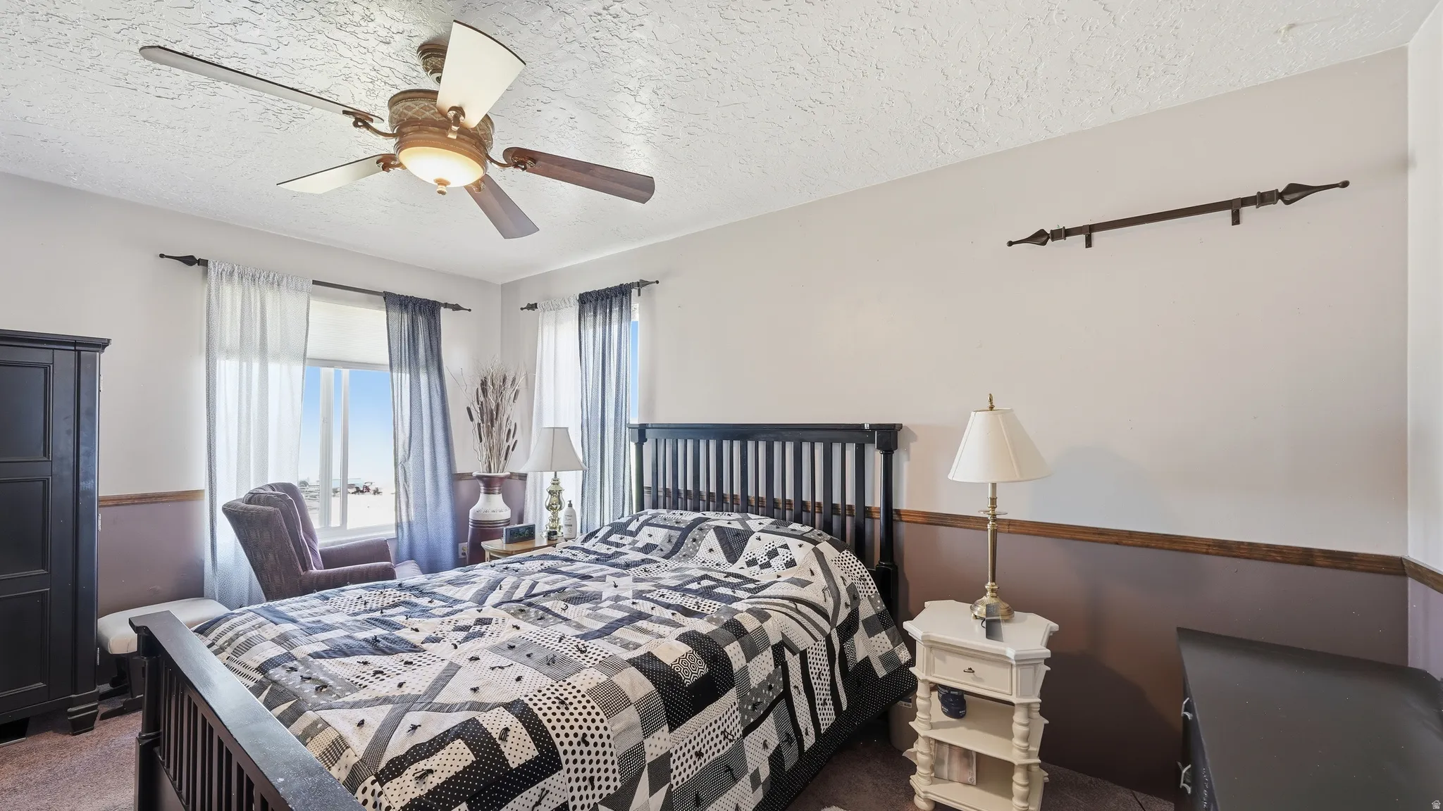 Spacious bedroom featuring large windows providing natural light, a ceiling fan with integrated lighting, and a two-tone wall finish with a decorative wood accent rail