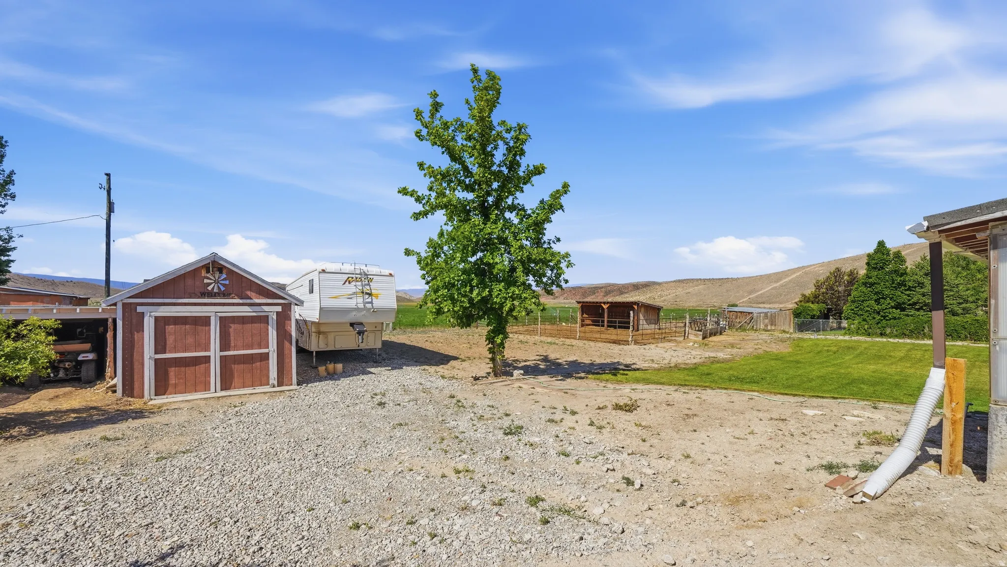 Expansive gravel yard featuring a red storage shed with barn doors, a mature deciduous tree, a grassy area, and a wood-framed shelter in the background