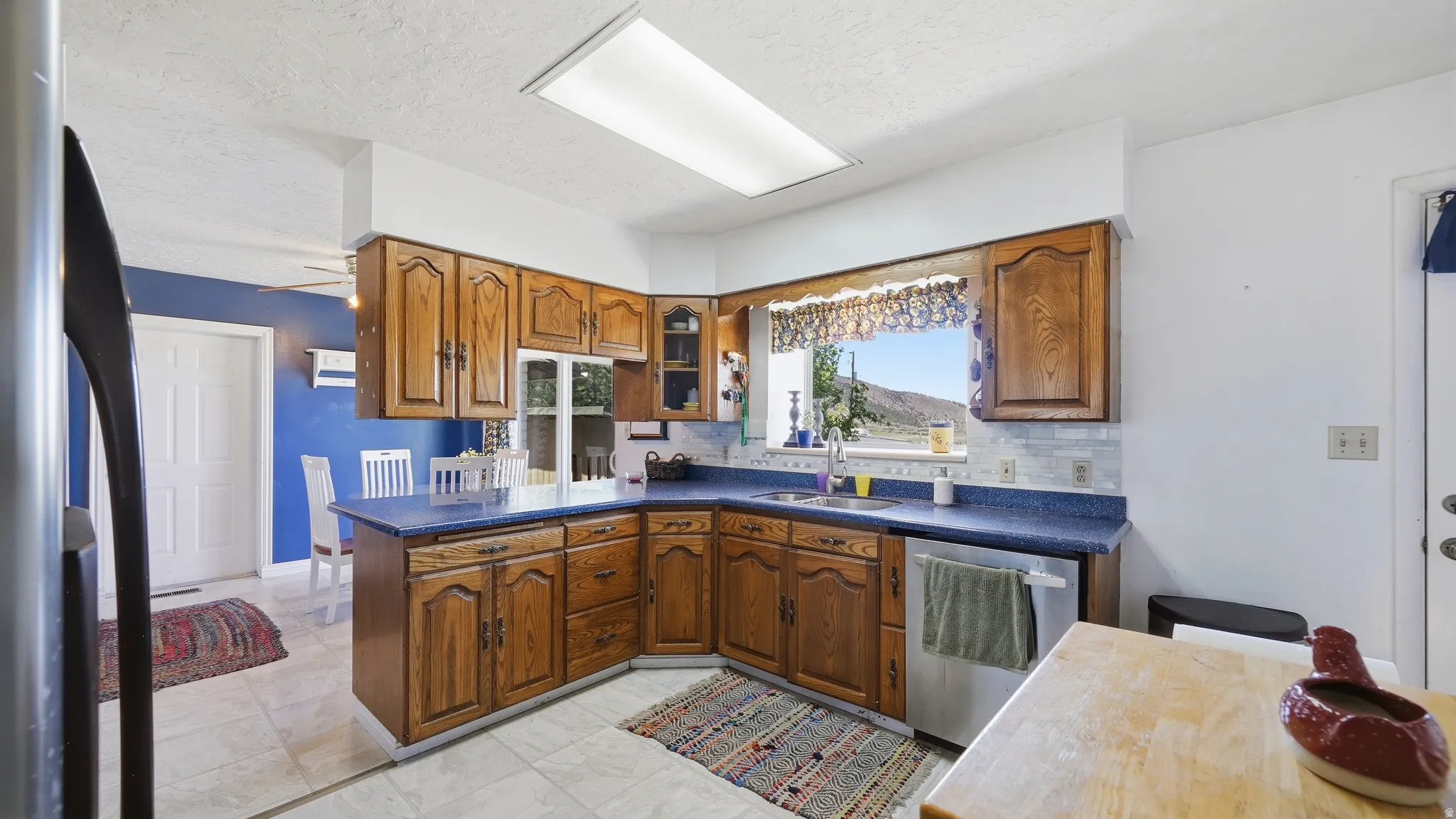 Kitchen featuring wood cabinetry, blue countertops, tile flooring, a built-in dishwasher, and a window with outdoor scenery