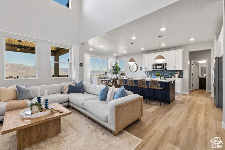 Spacious open-concept living area featuring high ceilings, wood-finish flooring, a large kitchen island with pendant lighting, white cabinetry, and a blue tile backsplash