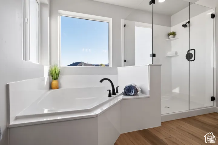 Corner soaking tub with a black faucet, glass-enclosed shower, white tile shower surround, and wood-finish flooring