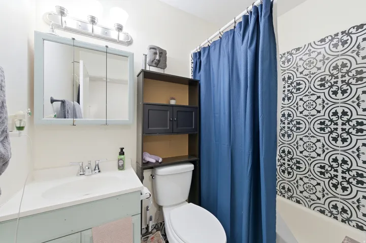 Full bathroom featuring a vanity with an integrated sink, a medicine cabinet with a light-colored frame, a three-light vanity fixture, patterned shower wall tile, and a built-in storage shelving unit