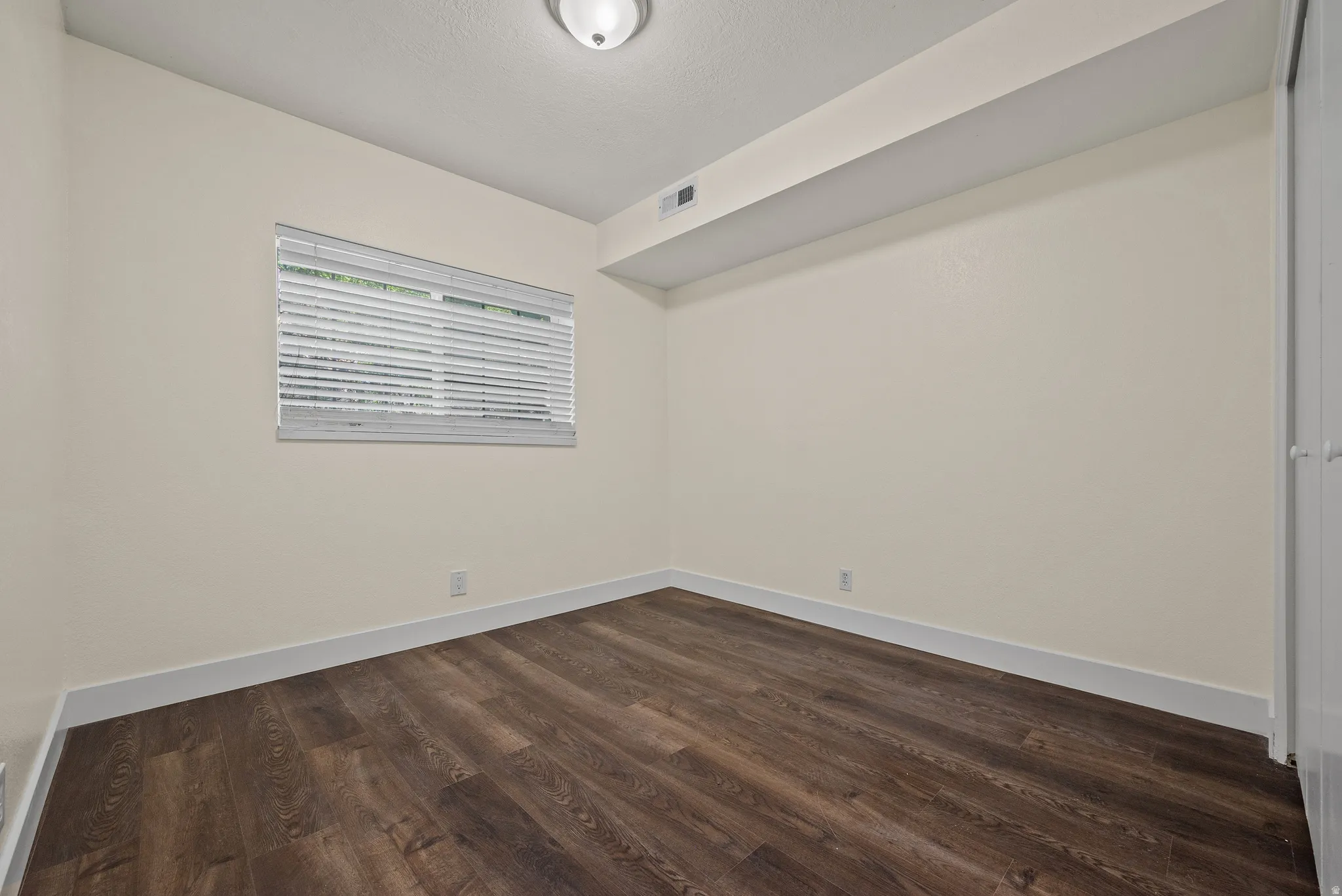 Interior room featuring wood-finish flooring, light-toned walls, a single window with horizontal blinds, and a flush-mount ceiling light fixture
