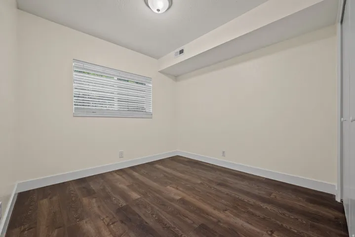 Interior room featuring wood-finish flooring, light-toned walls, a single window with horizontal blinds, and a flush-mount ceiling light fixture