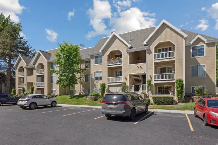 Multi-story residential building featuring light-colored siding, multiple balconies with white railings, and a large paved parking area