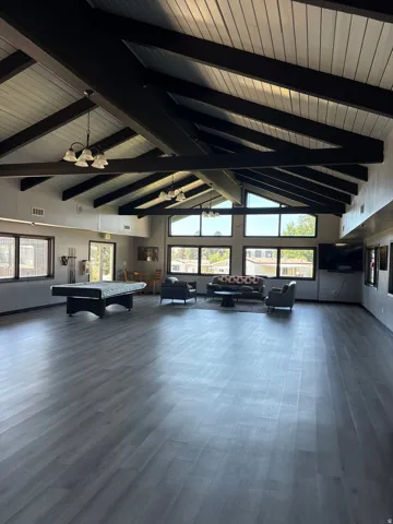 Spacious common area featuring vaulted ceilings with exposed beams, wood-finish flooring, abundant natural light from large windows, and multiple pendant light fixtures