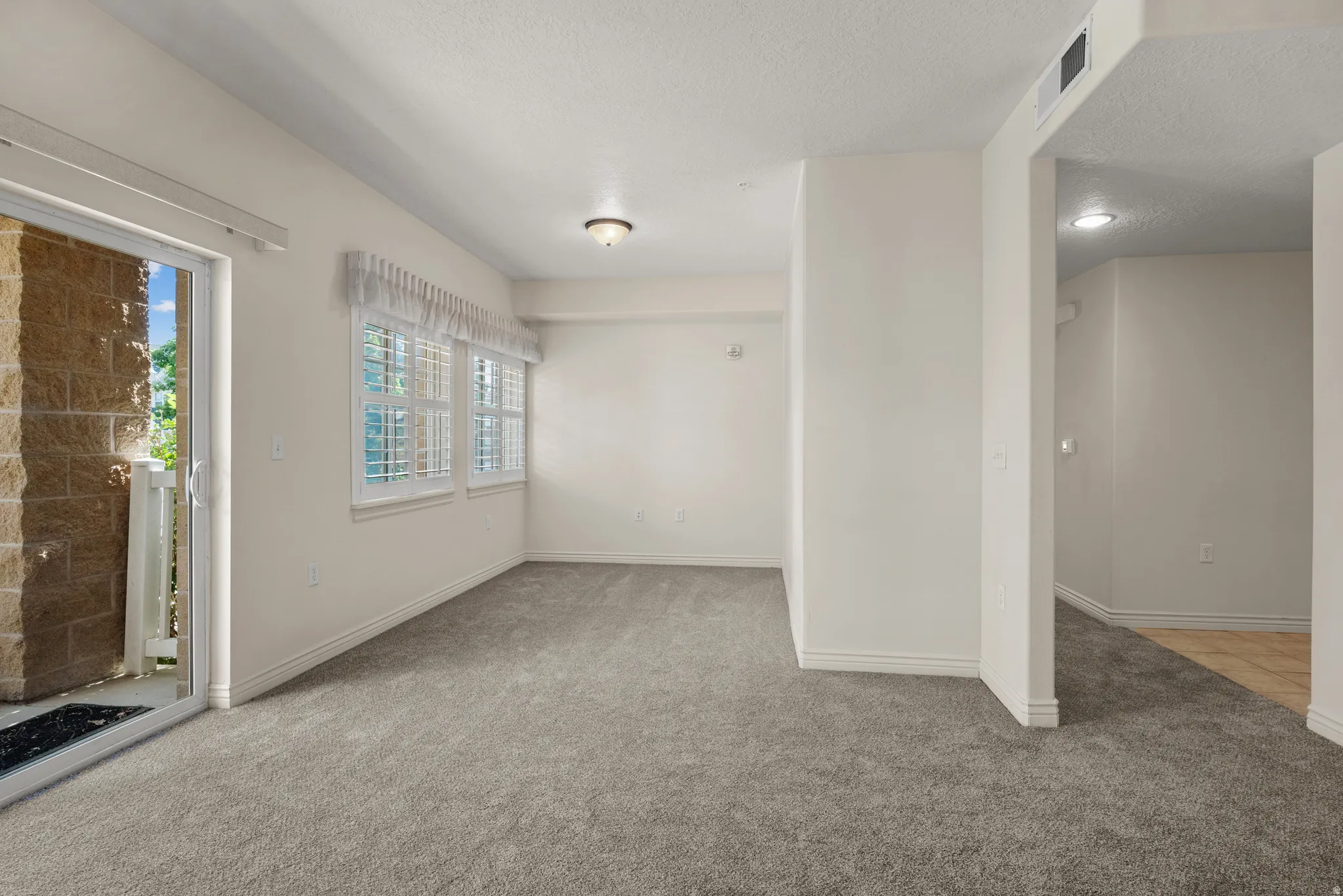 Carpeted living area featuring a sliding glass door to a private patio, three windows with white plantation shutters, and a semi-flush mount ceiling light