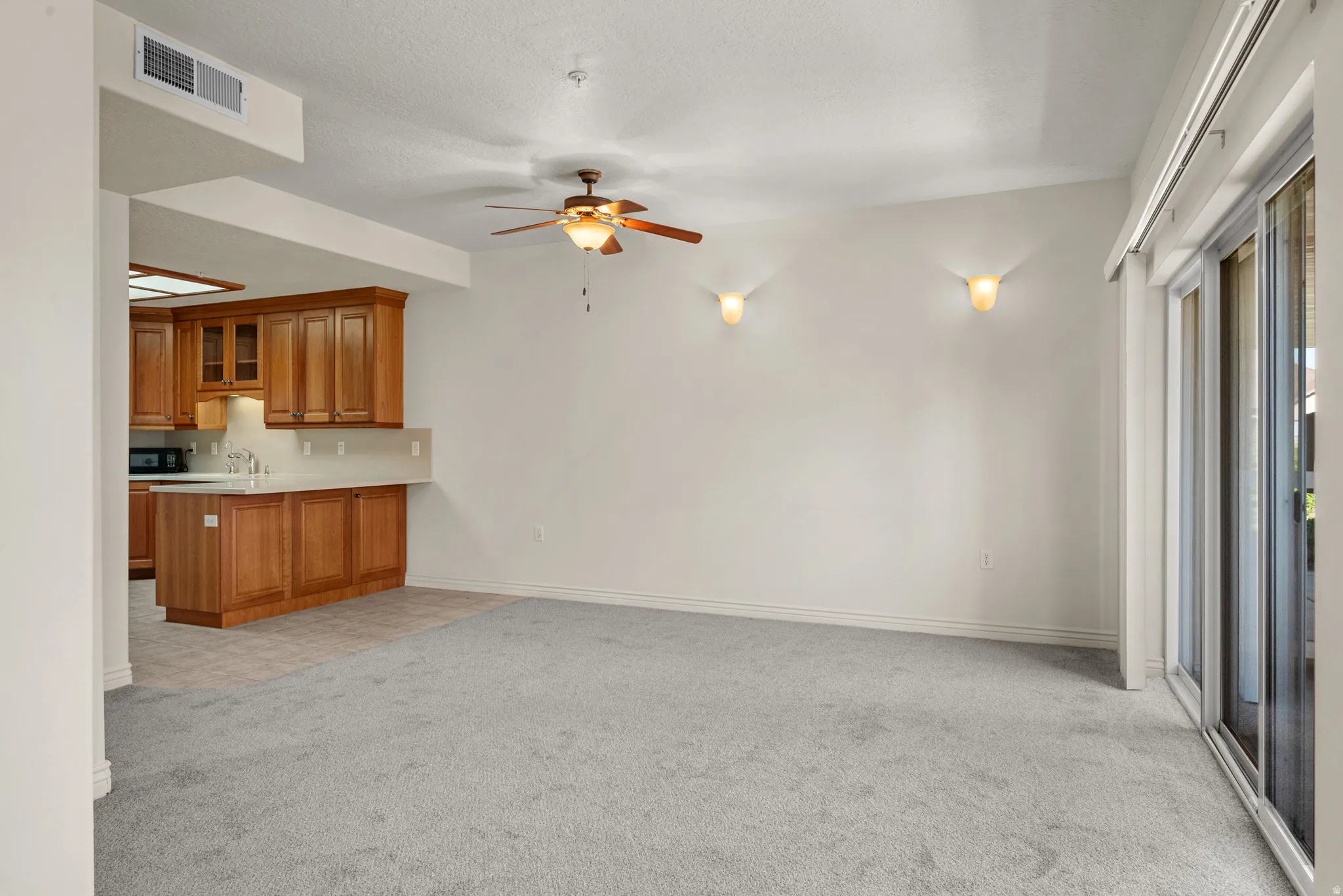 Expansive carpeted living area featuring a ceiling fan with integrated lighting, two wall sconces, and sliding glass doors