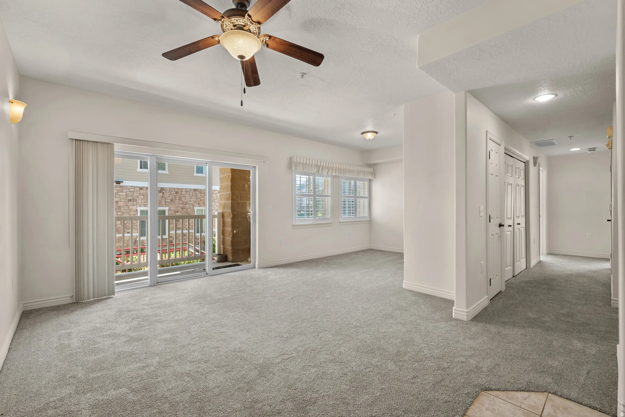 Spacious living area featuring light gray carpeting, a ceiling fan with integrated lighting, and a sliding glass door leading to a private balcony