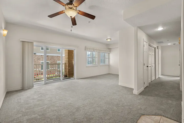 Spacious living area featuring light gray carpeting, a ceiling fan with integrated lighting, and a sliding glass door leading to a private balcony