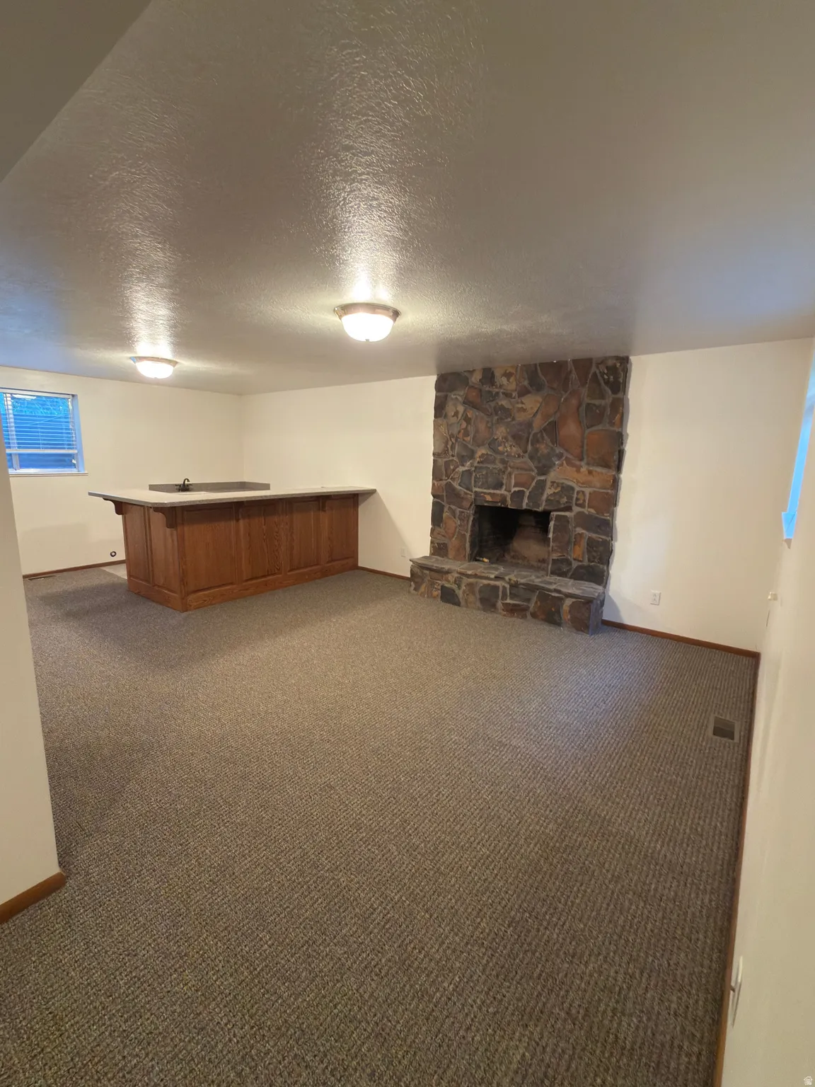 Spacious room featuring a stone fireplace, built-in bar with a sink, textured carpet flooring, and two flush mount ceiling lights