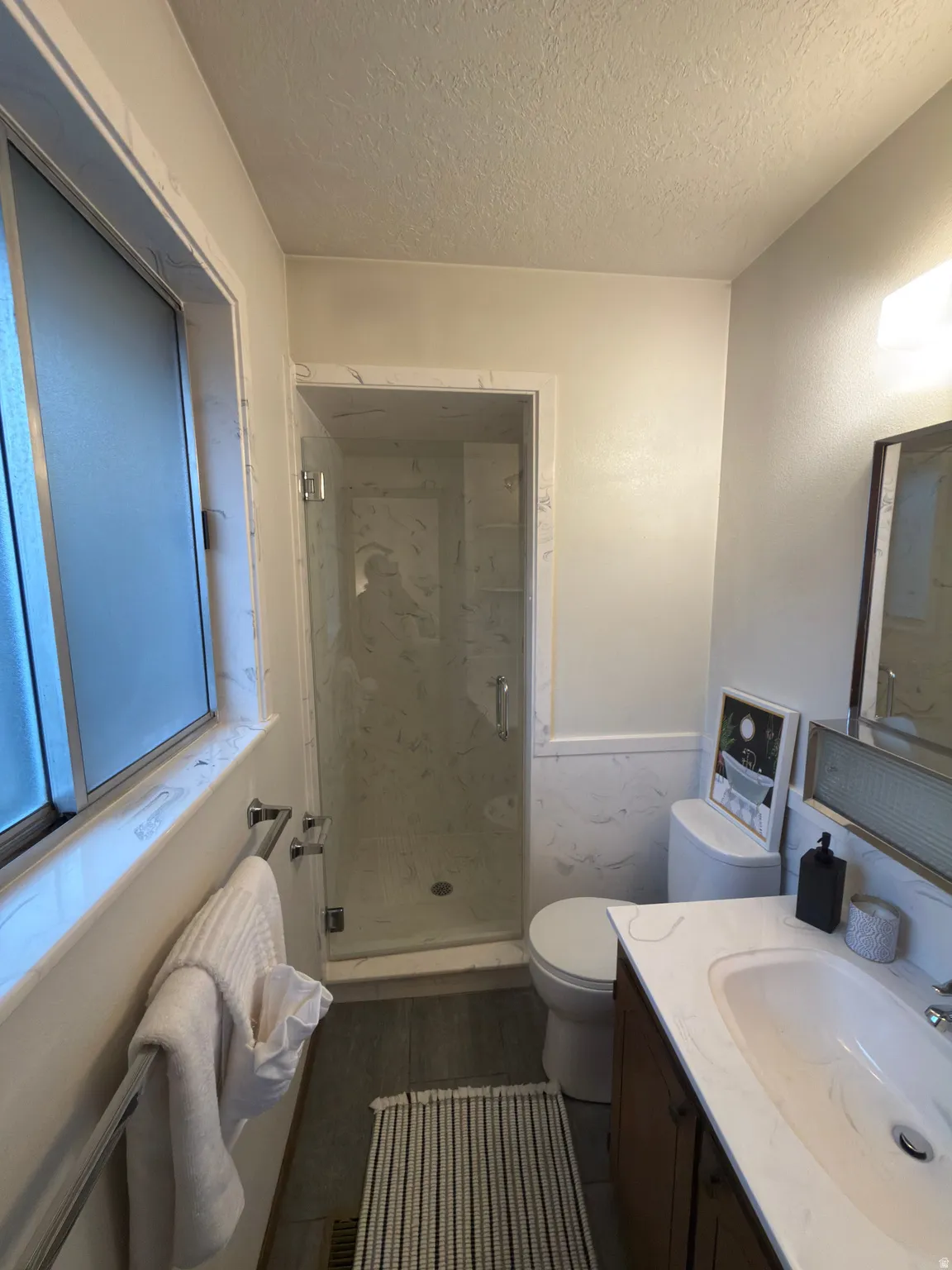 Bathroom featuring a glass-enclosed standing shower with white-patterned wall panels, a wood-finish vanity with an integrated sink and white countertop, and a frosted window