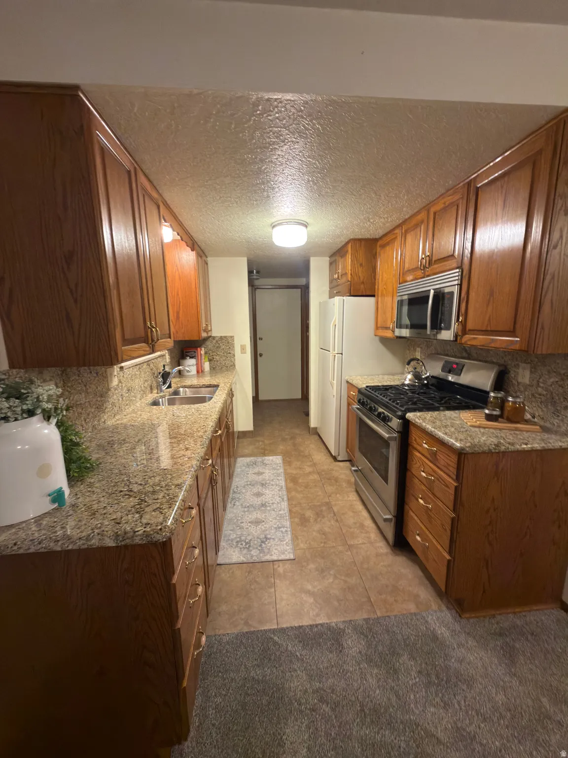 Galley kitchen featuring wood-finish cabinetry, stone-finish countertops, stainless steel appliances including a range and microwave, and tile flooring