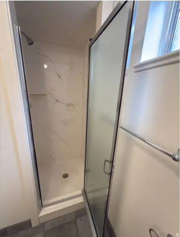 Walk-in shower with a glass door, white shower surround, integrated shelving, and a chrome showerhead
