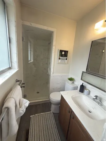 Bathroom featuring a glass-enclosed shower with white marble-look paneling, a wood-finish vanity with an integrated sink and white marble-look countertop, dark gray wood-finish flooring, and a window with frosted glass