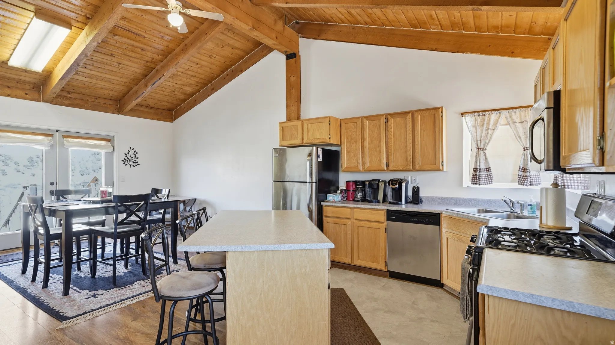 Kitchen and dining area featuring a vaulted wood-plank ceiling with exposed beams, light wood-finish cabinetry, stainless steel appliances, and a central island with bar seating