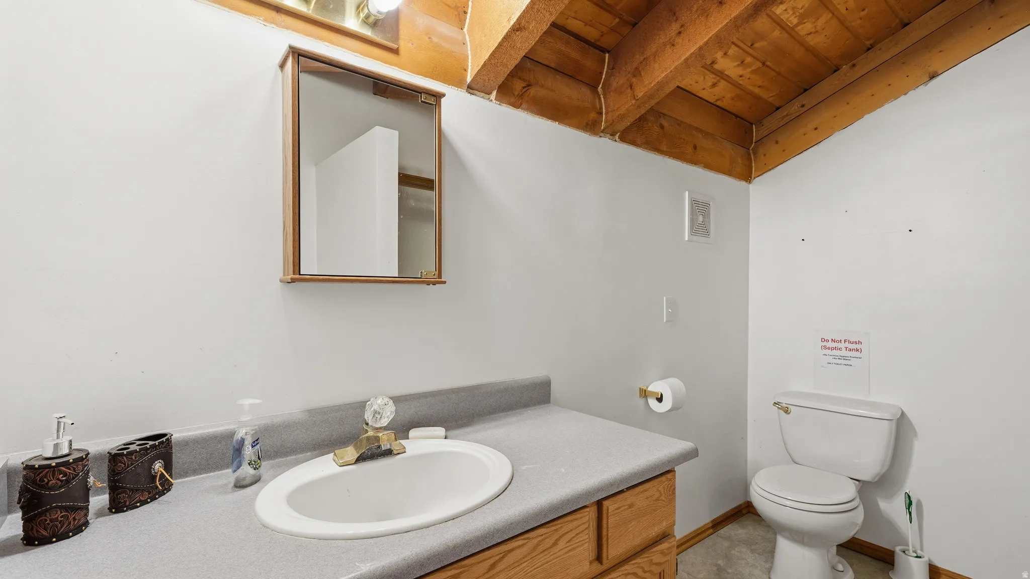 Bathroom featuring exposed wood ceiling beams, a wood-finish vanity with a built-in sink, a wall-mounted medicine cabinet, a toilet, and a vinyl flooring
