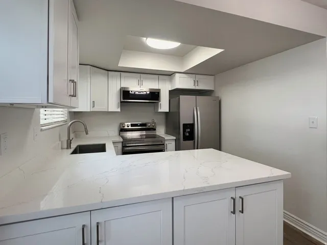 Modern kitchen featuring white shaker cabinetry with brushed nickel hardware, light-toned countertops with subtle veining, stainless steel appliances including a refrigerator and range, and an integrated kitchen sink with a gooseneck faucet
