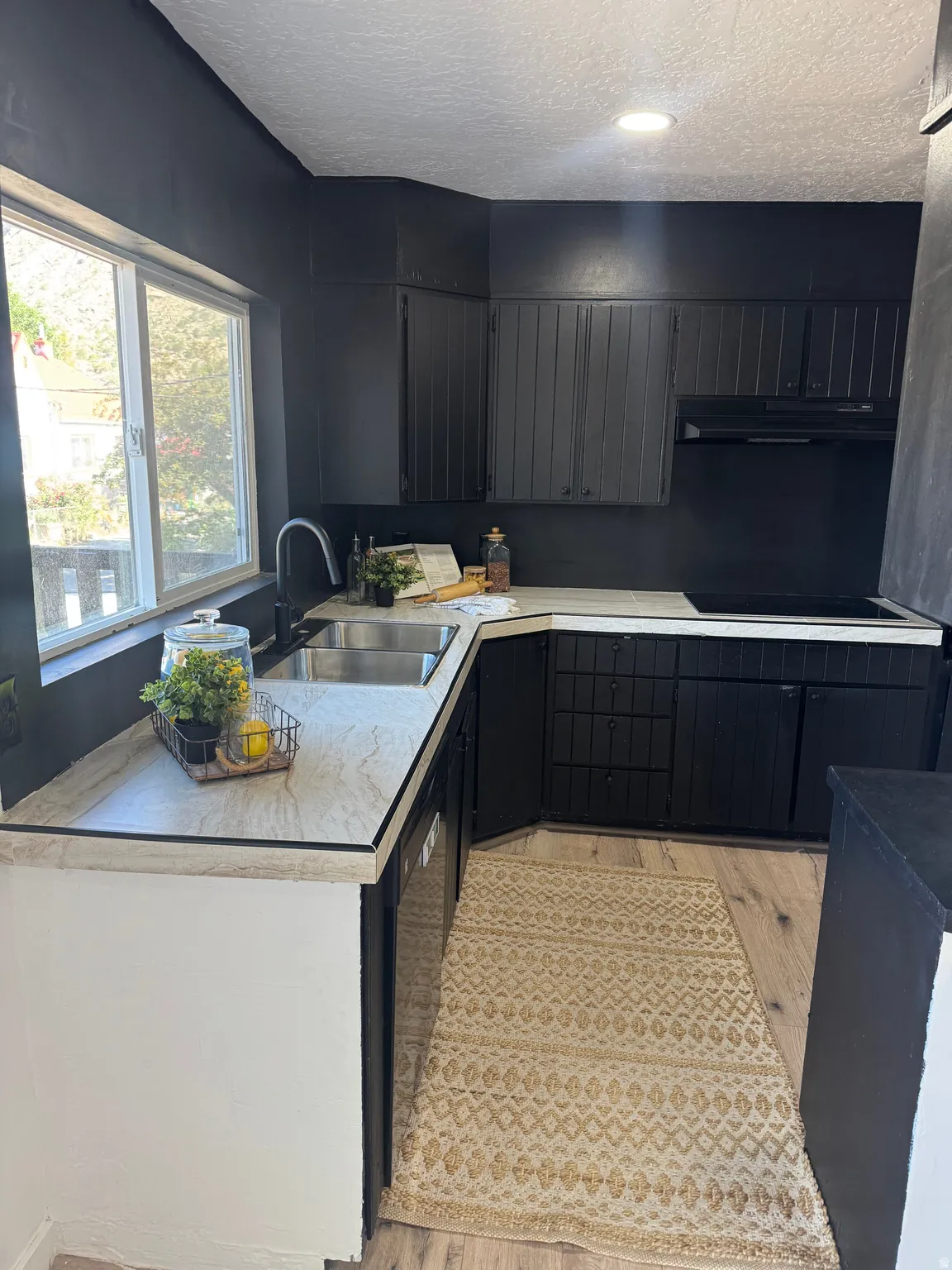 Contemporary kitchen featuring black cabinetry, light-toned countertops, a stainless steel double basin sink, and wood-finish flooring