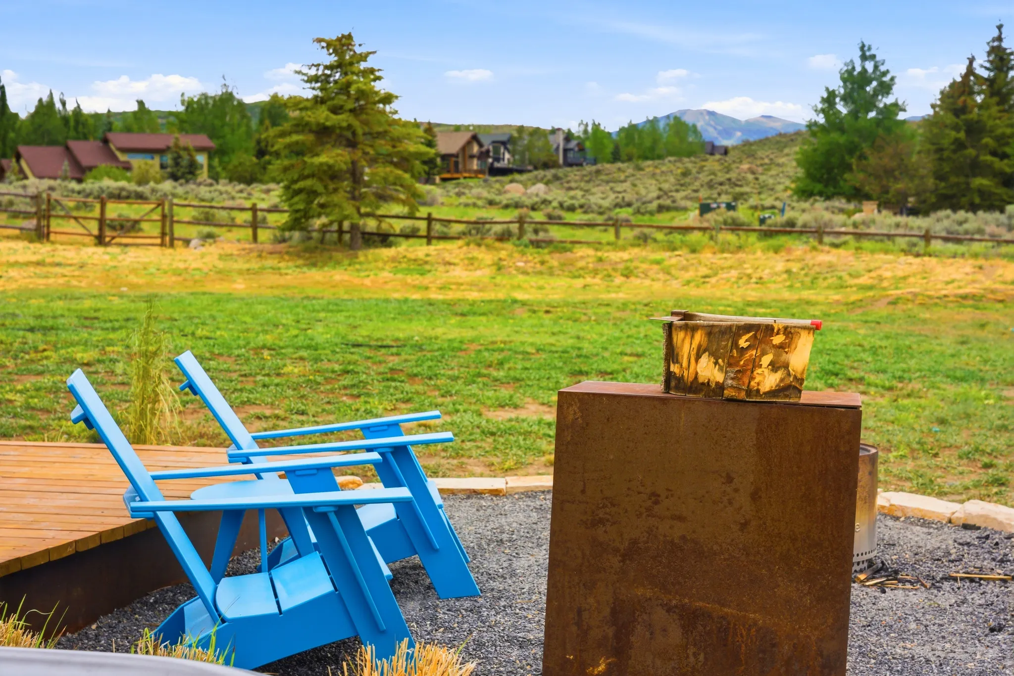 Outdoor living space featuring a wood deck, gravel patio area, a metal fire pit structure, and an expansive grassy field bordered by a wooden fence