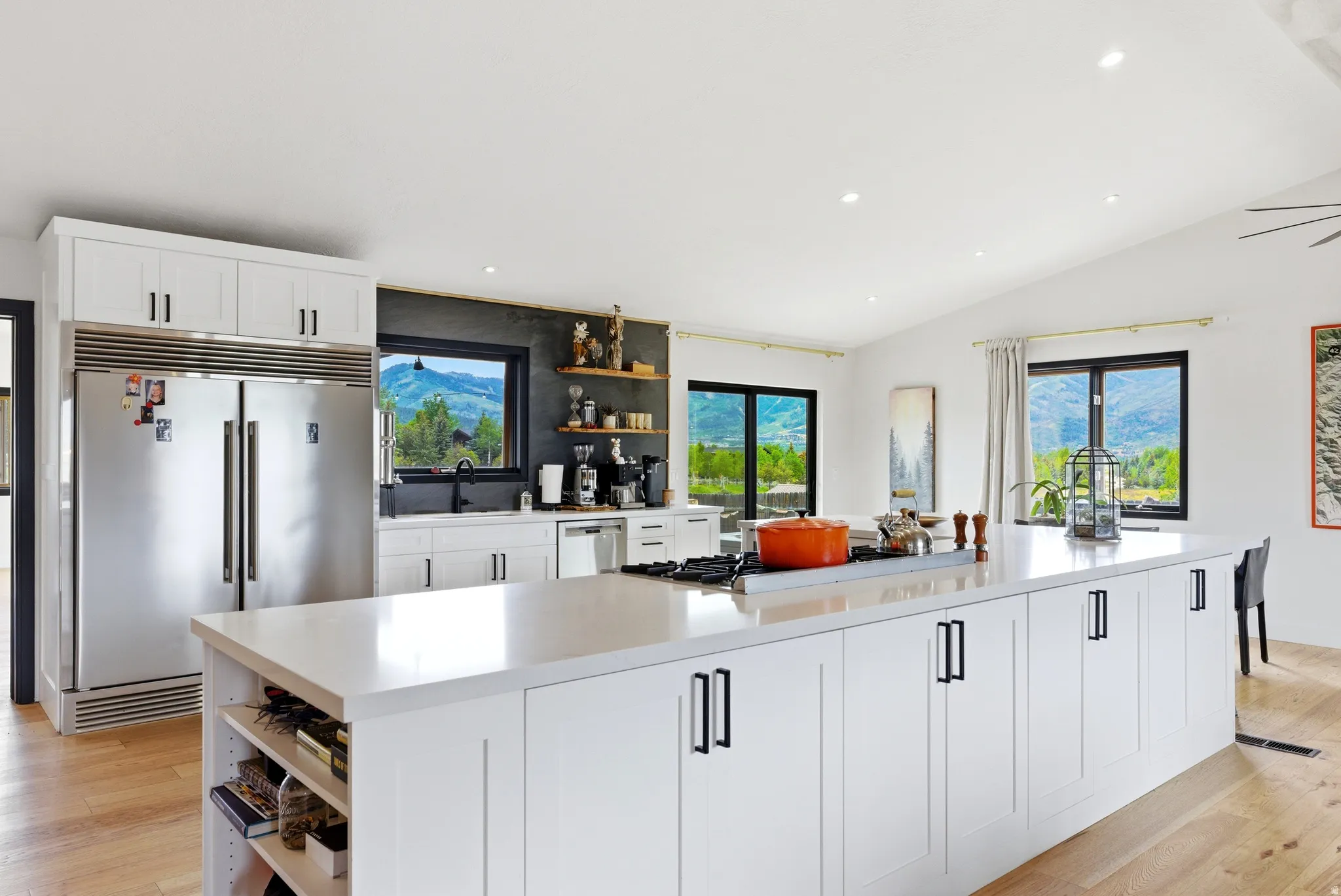 Spacious kitchen featuring a large white island with integrated cooktop, light wood-finish flooring, white cabinetry with black hardware, recessed lighting, and large windows providing natural light