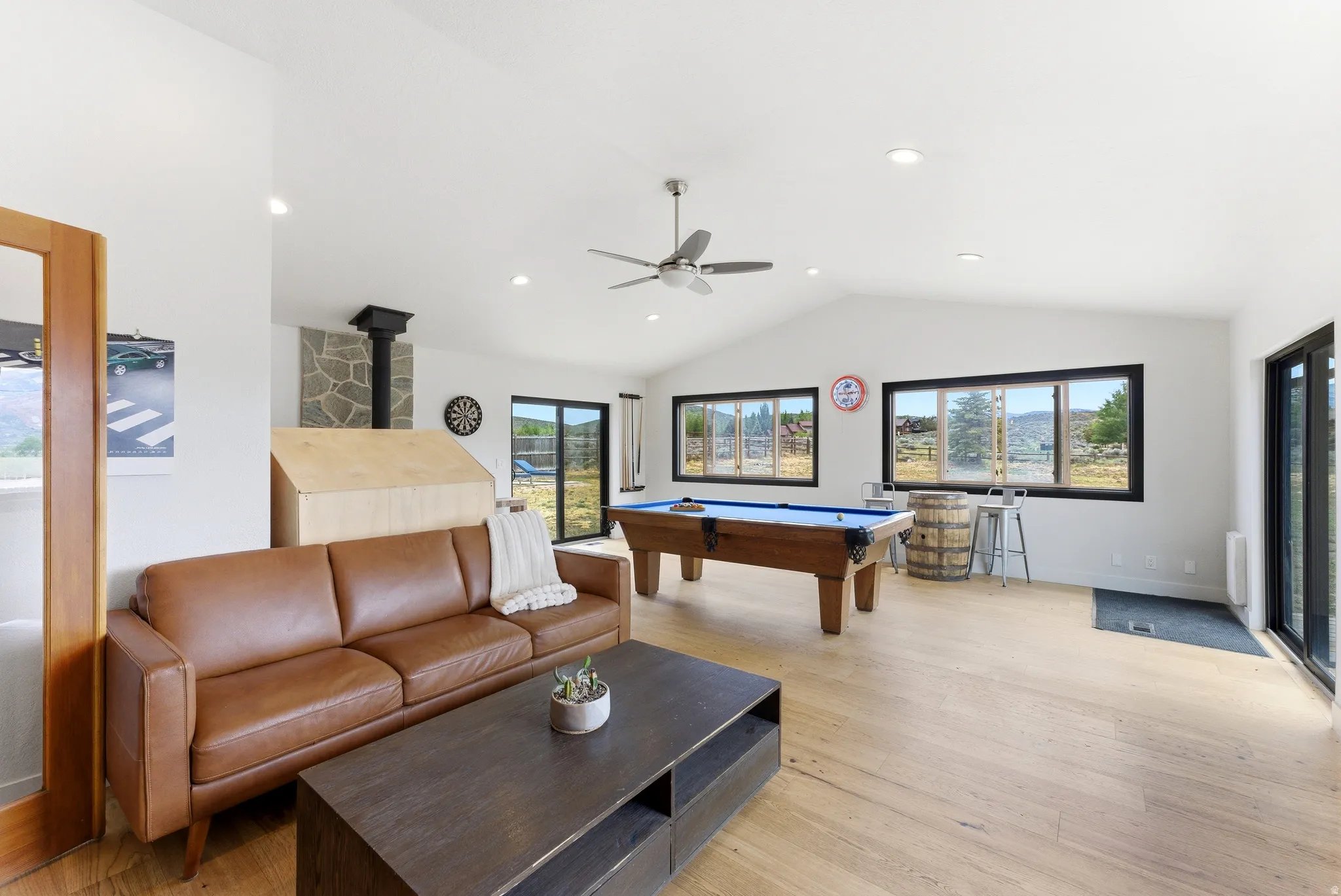 Spacious room featuring vaulted ceilings, light wood-finish flooring, recessed lighting, and a prominent stone-clad fireplace with a black stovepipe