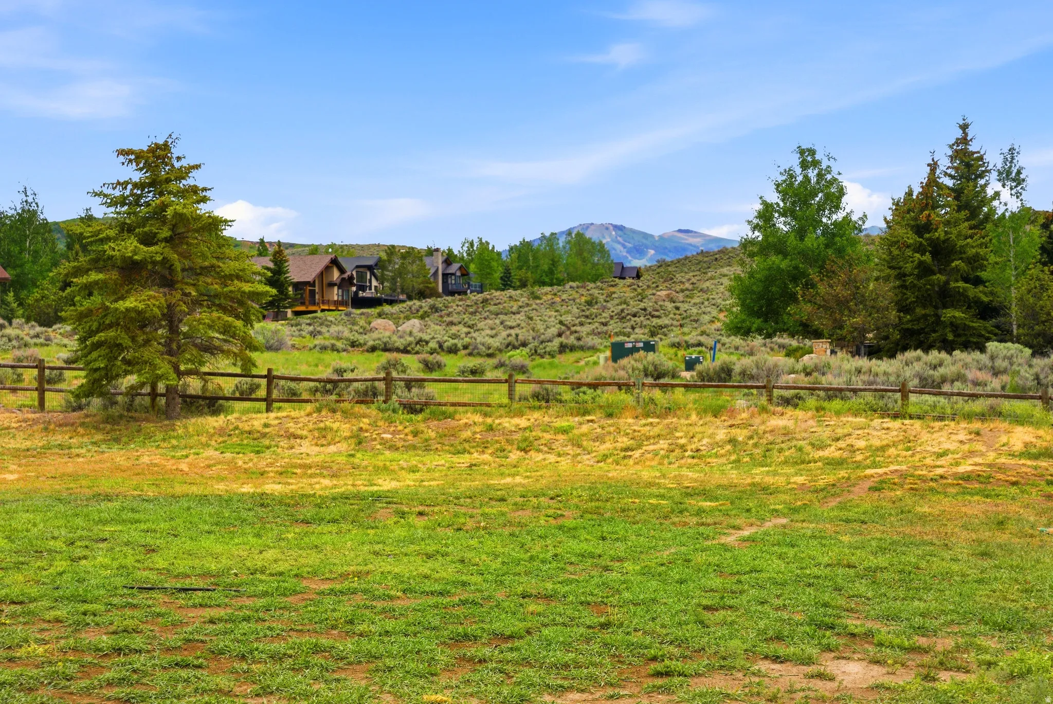 Expansive green space featuring a split-rail fence, mature evergreen trees, and distant mountain scenery