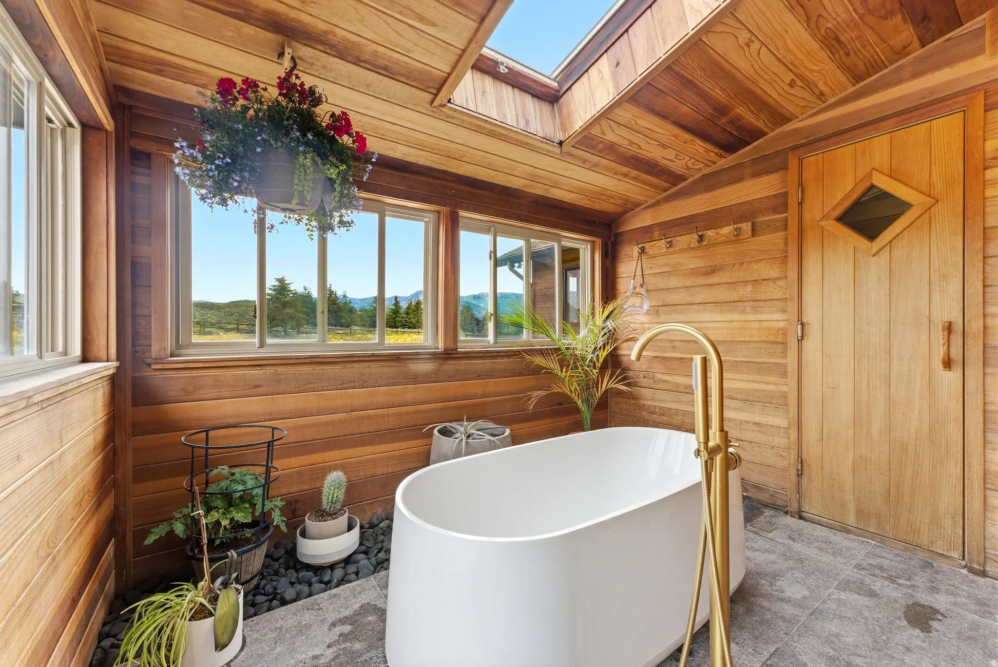 Freestanding soaking tub with a brass floor-mounted faucet, surrounded by wood-paneled walls and ceiling, a skylight, and large windows revealing a mountain backdrop