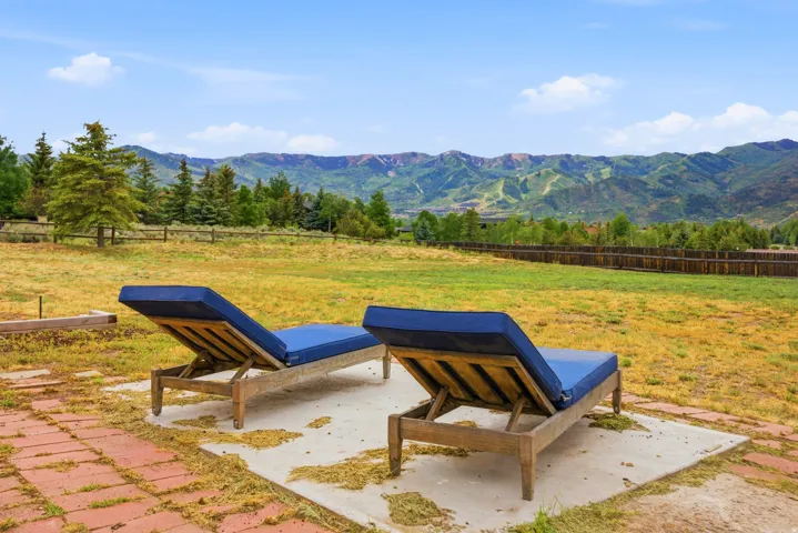 Expansive outdoor area featuring a large grassy lawn, natural wood perimeter fencing, mature evergreen trees, and distant mountainous terrain