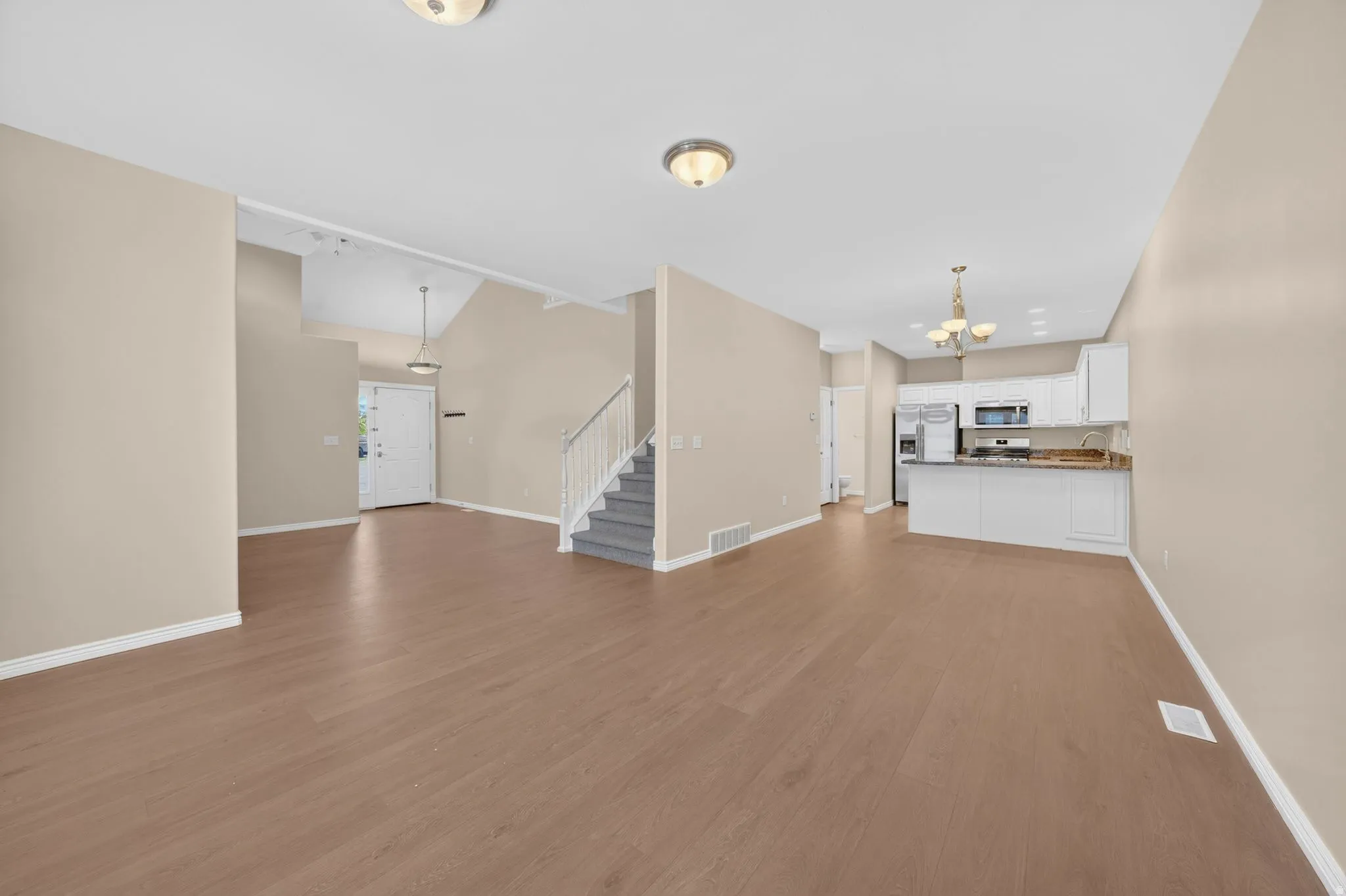 Open-concept living area featuring wood-finish flooring, a kitchen with white cabinetry and stainless steel appliances, a staircase with carpeting, a prominent entry door, and recessed lighting fixtures
