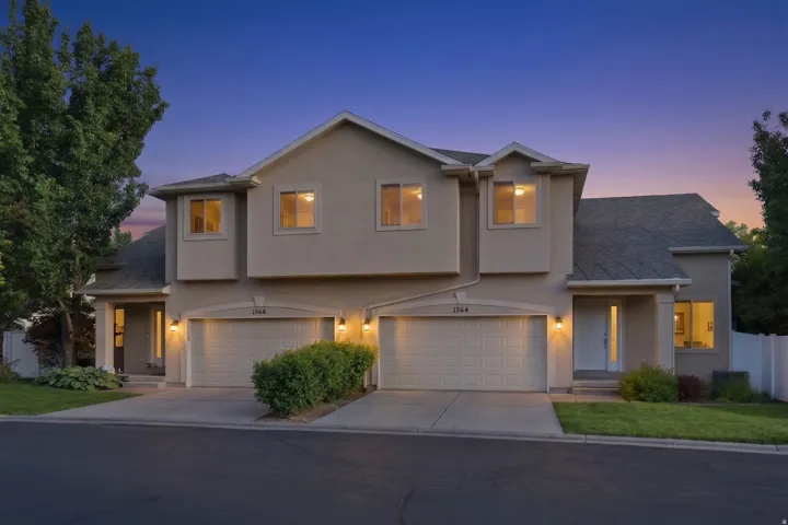 Contemporary stucco exterior featuring a gabled roofline, multiple windows, attached garages with individual driveways, and integrated outdoor lighting